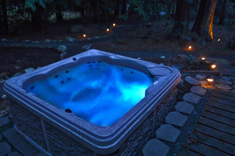 The hot tub at dusk, lit blue from inside, with small path lights glowing along a stone walkway and the forest behind.